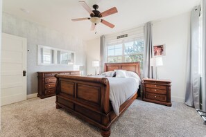 Een slaapkamer met een houten bed, een plafondventilator, een nachtkastje, een dressoir, een spiegel en een raam met gordijnen.