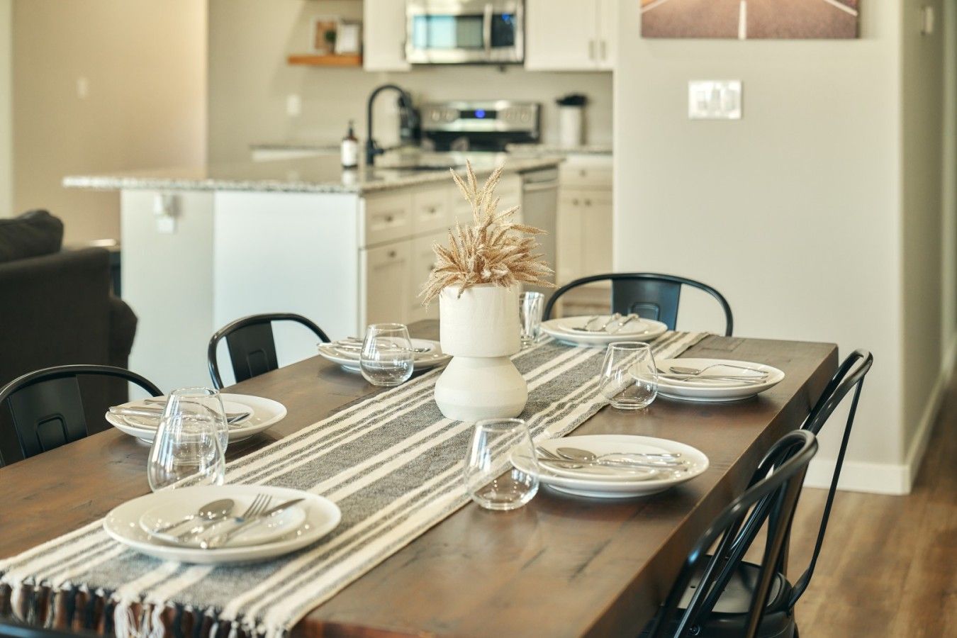 A dining table set with plates, glasses, and cutlery, a vase with dried plants, and a striped table runner.