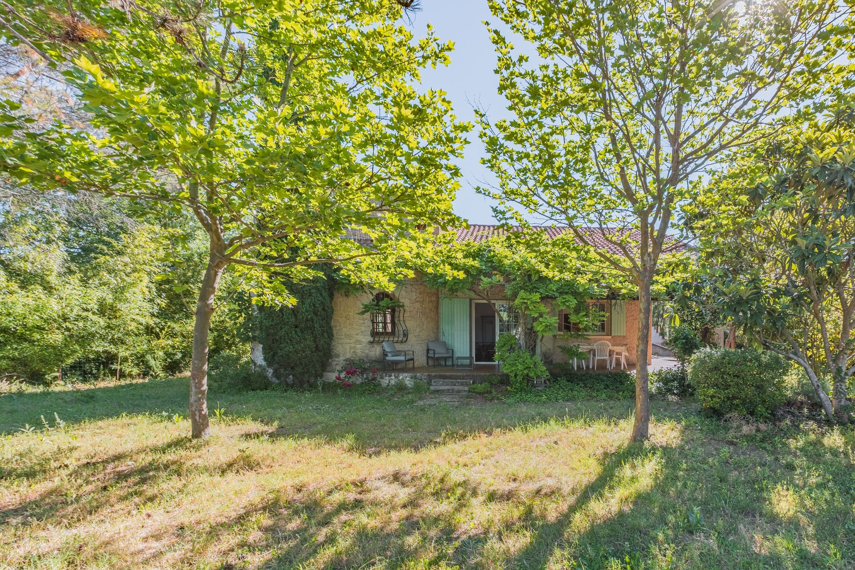 A rustic stone house with a porch, surrounded by lush greenery and trees.