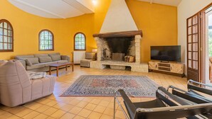 A living room with a stone fireplace, a sofa, a television, and a large window. - Le Mas des Trois Sources - Grand jardin (Gignac)