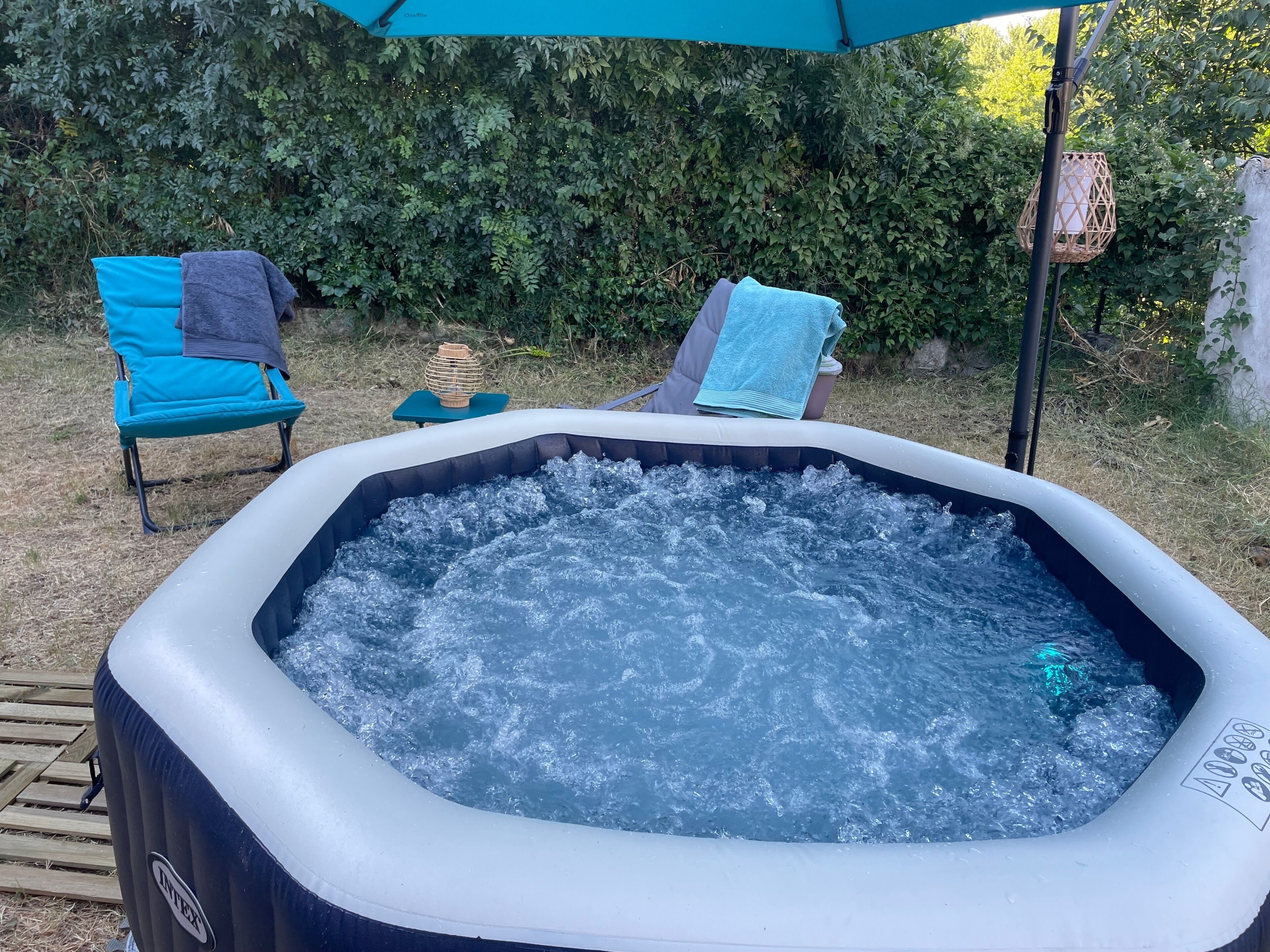 An inflatable hot tub with a wooden deck and chairs, surrounded by greenery.