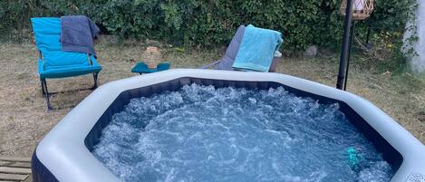 An inflatable hot tub with a wooden deck and chairs, surrounded by greenery.