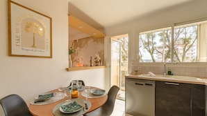 Un coin repas avec une table en bois dressée pour deux personnes, une cuisine équipée d’un lave-vaisselle et une grande fenêtre donnant sur les arbres.