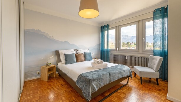 Une chambre à coucher comprenant un lit, une chaise, une table de chevet, une lampe, un radiateur et une fenêtre donnant sur les montagnes.