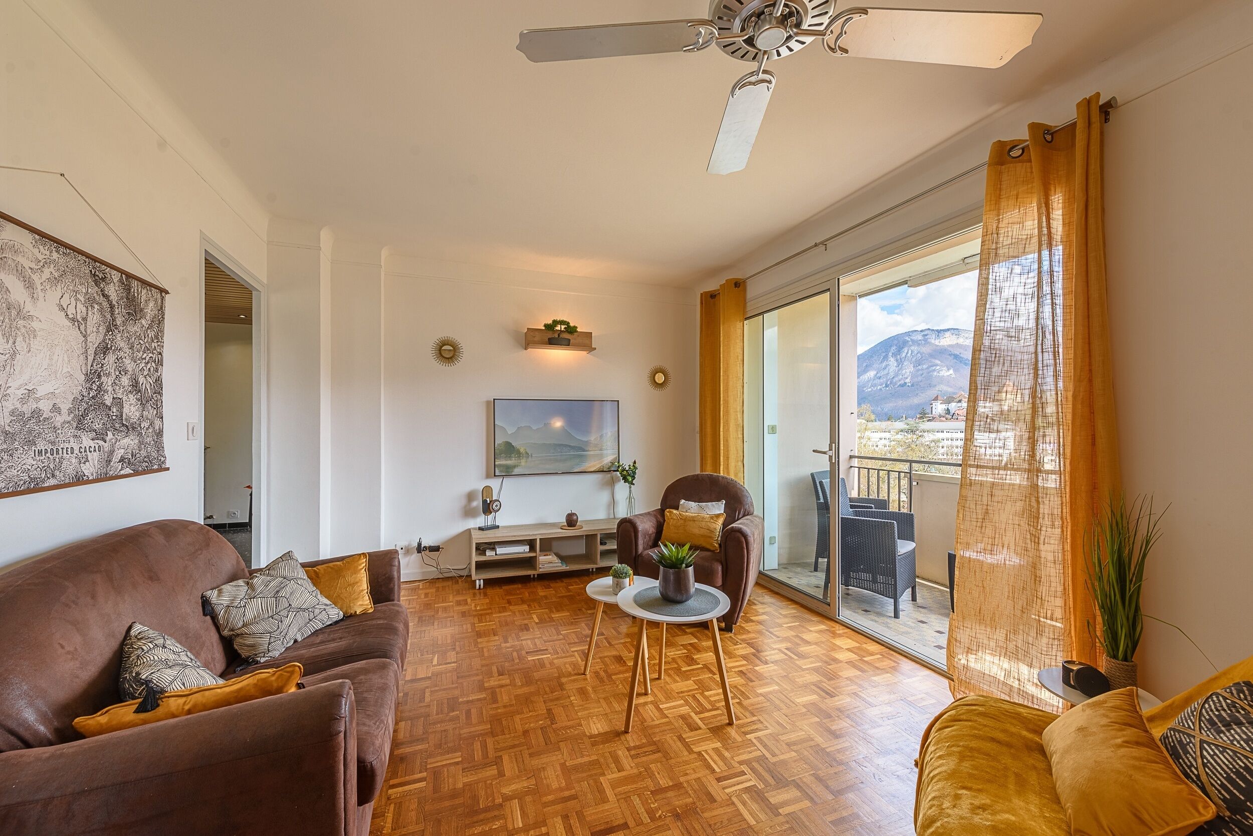 Un salon comprenant un canapé, un fauteuil, une table basse et un téléviseur. Il y a un balcon avec vue sur les montagnes.
