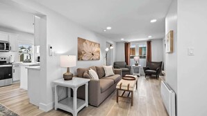 A modern living room with a sofa, a coffee table, and a lamp. There is a kitchen area with cabinets and appliances. - Spacious 3 Bdrm Haven - Steps to Downtown Ch'town (Charlottetown)