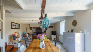 Un comedor con una mesa de madera, sillas, un florero con flores, lámparas colgantes y una cocina al fondo.