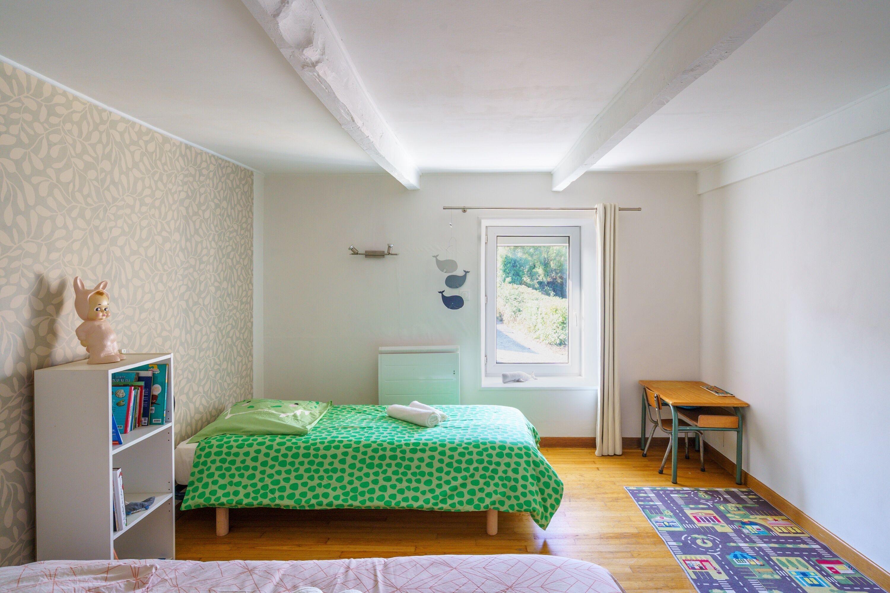 A bedroom with a bed, a desk, a bookshelf, and a window with a view of greenery.