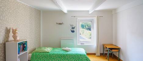A bedroom with a bed, a desk, a bookshelf, and a window with a view of greenery.