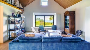 Una habitación amplia con techo alto, un ventanal que ofrece vistas a zonas verdes, un sofá azul, una mesa de centro blanca y una estantería.
