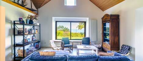 Un'ampia stanza con soffitto alto, una grande finestra che si affaccia sul verde, un divano blu, un tavolino da caffè bianco e una libreria.