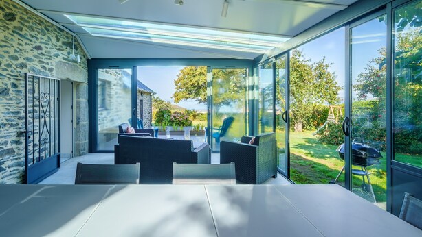Un área de comedor moderna al aire libre con cerramiento de vidrio, paredes de piedra y vistas a un jardín.