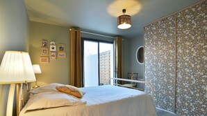 A bedroom with a bed, bedside lamps, a window with curtains, a wall-mounted shelf, and a floral-patterned partition. - Le Petit Carrelet - 10min de Dinan (Saint-Samson-sur-Rance)