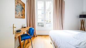 A hotel room with a bed, a desk with a chair, a lamp, and a window with curtains. - Grands Boulevards Home (Paris)