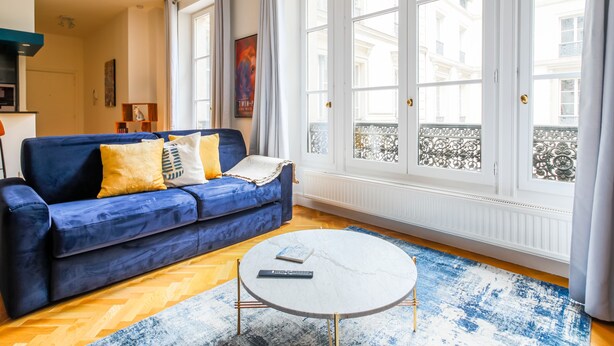 A living room with a blue sofa, a round coffee table, and large windows with a view of buildings. - Grands Boulevards Home (Paris)