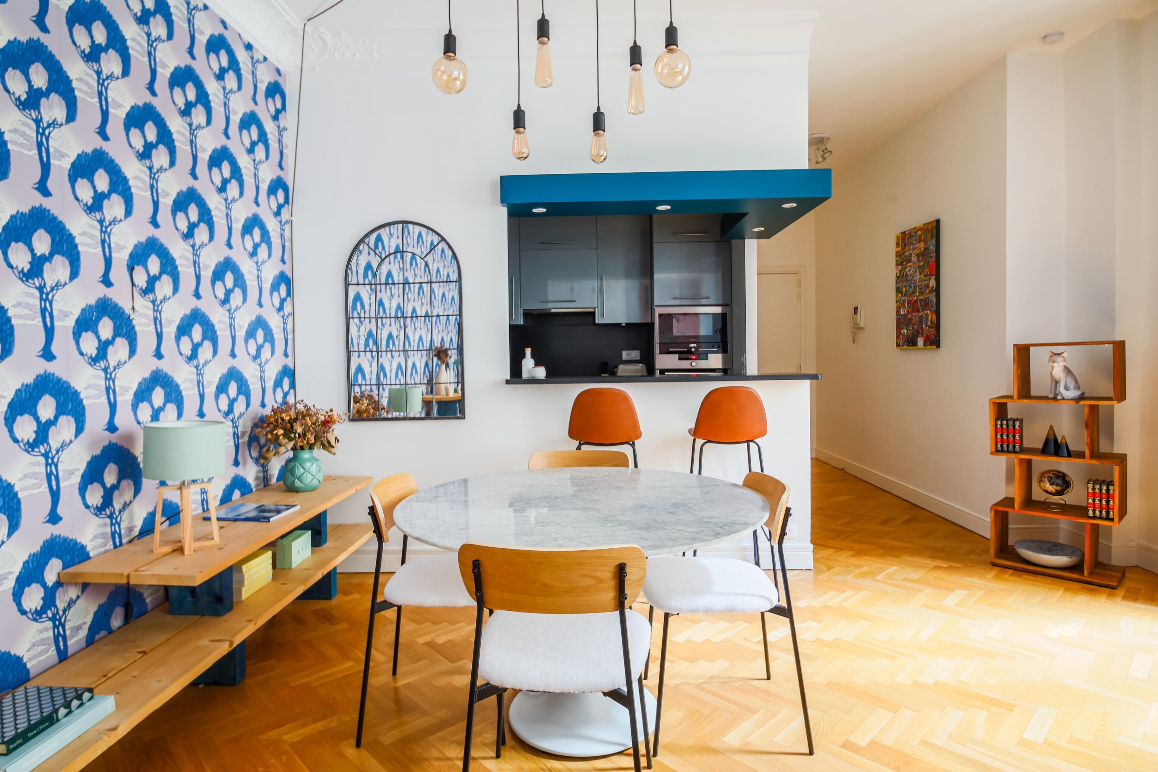 Een eetgedeelte met een ronde tafel, stoelen, een bank, een muur met een blauw patroon, een keuken op de achtergrond en een boekenkast met decoratieve objecten.