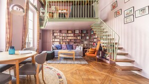 A cozy living room with a wooden herringbone floor, a sofa, a coffee table, a bookshelf, and a staircase with a green railing. - Saint Michel Seine Loft (Paris)