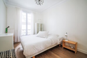 Een slaapkamer met een bed, een nachtkastje met een lamp, een radiator, een raam met gordijnen, en een witte inbouwkast.