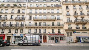 Ein mehrstöckiges Gebäude mit Balkonen, einem parkenden Lieferwagen und Geschäften, darunter eine Diner und ein Videospielgeschäft.
