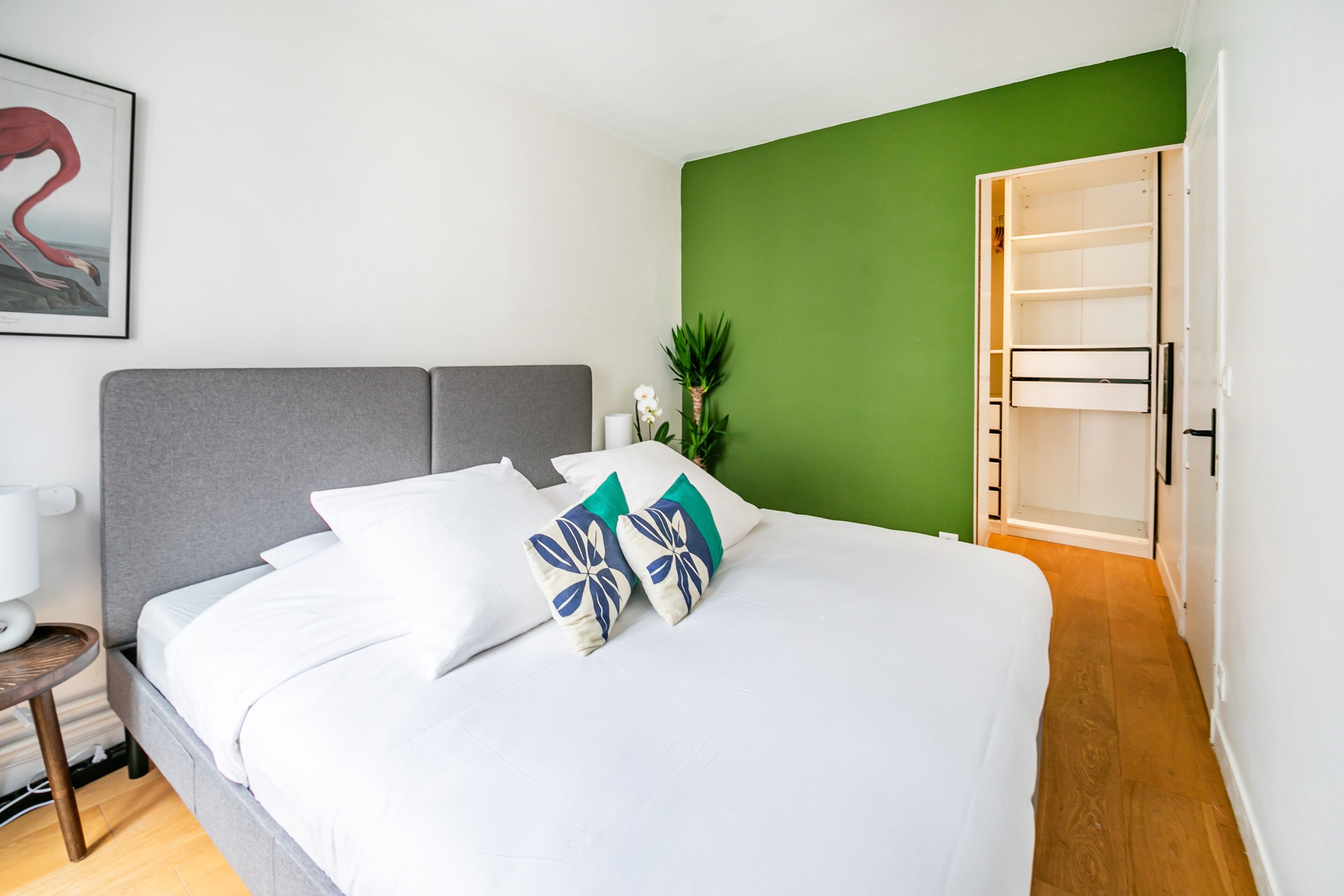 A neatly made bed with white linens and decorative pillows, a grey headboard, a green accent wall, and a wooden floor.
