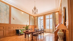 A spacious room with a large wooden dining table, chairs, a chandelier, and a large window with curtains.
