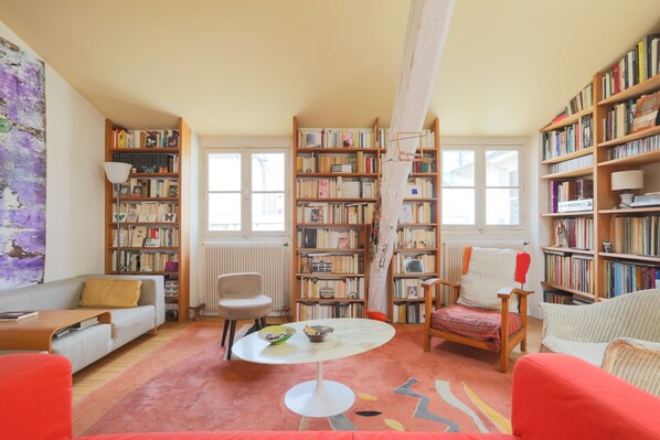 Ein gemütliches Wohnzimmer mit einer Couch, Sesseln, einem Couchtisch und einem Bücherregal voller Bücher.
