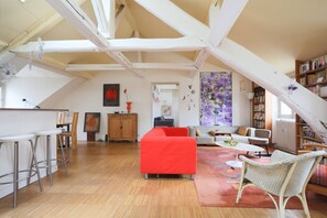 Ein Loft-Wohnraum mit freiliegenden Balken, einem roten Sofa, einem Essbereich mit Barhocker und einem Bücherregal.