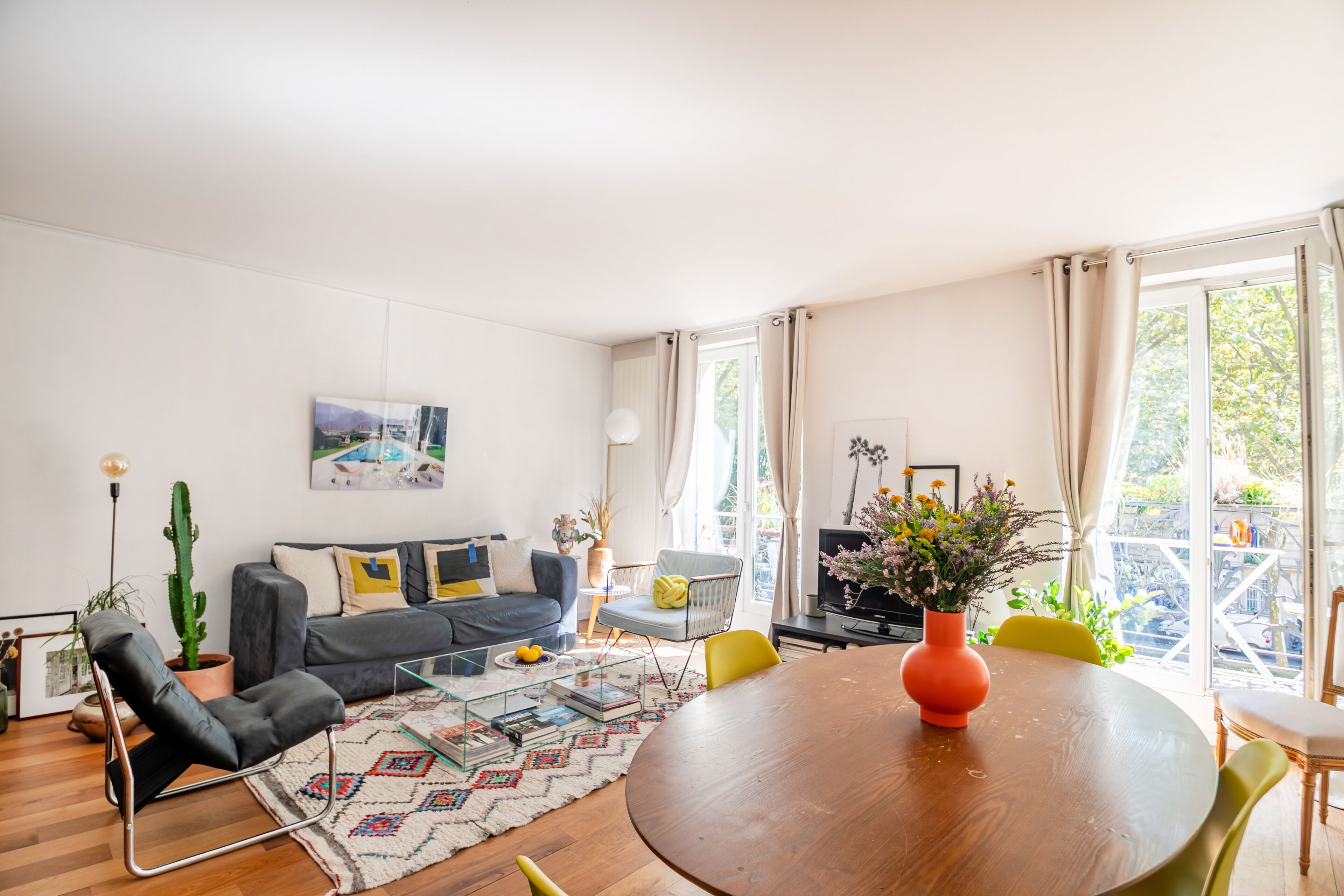 Un salon moderne avec une table ronde en bois, un canapé gris, une plante en pot (un cactus) et une grande fenêtre avec des rideaux.