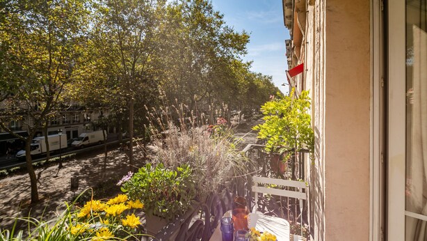 Un balcón con plantas en macetas, una mesa con un florero y una ventana con vista a la calle.