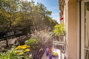 Een balkon met potplanten, een tafel met een vaas en een raam dat uitkijkt op een straat.