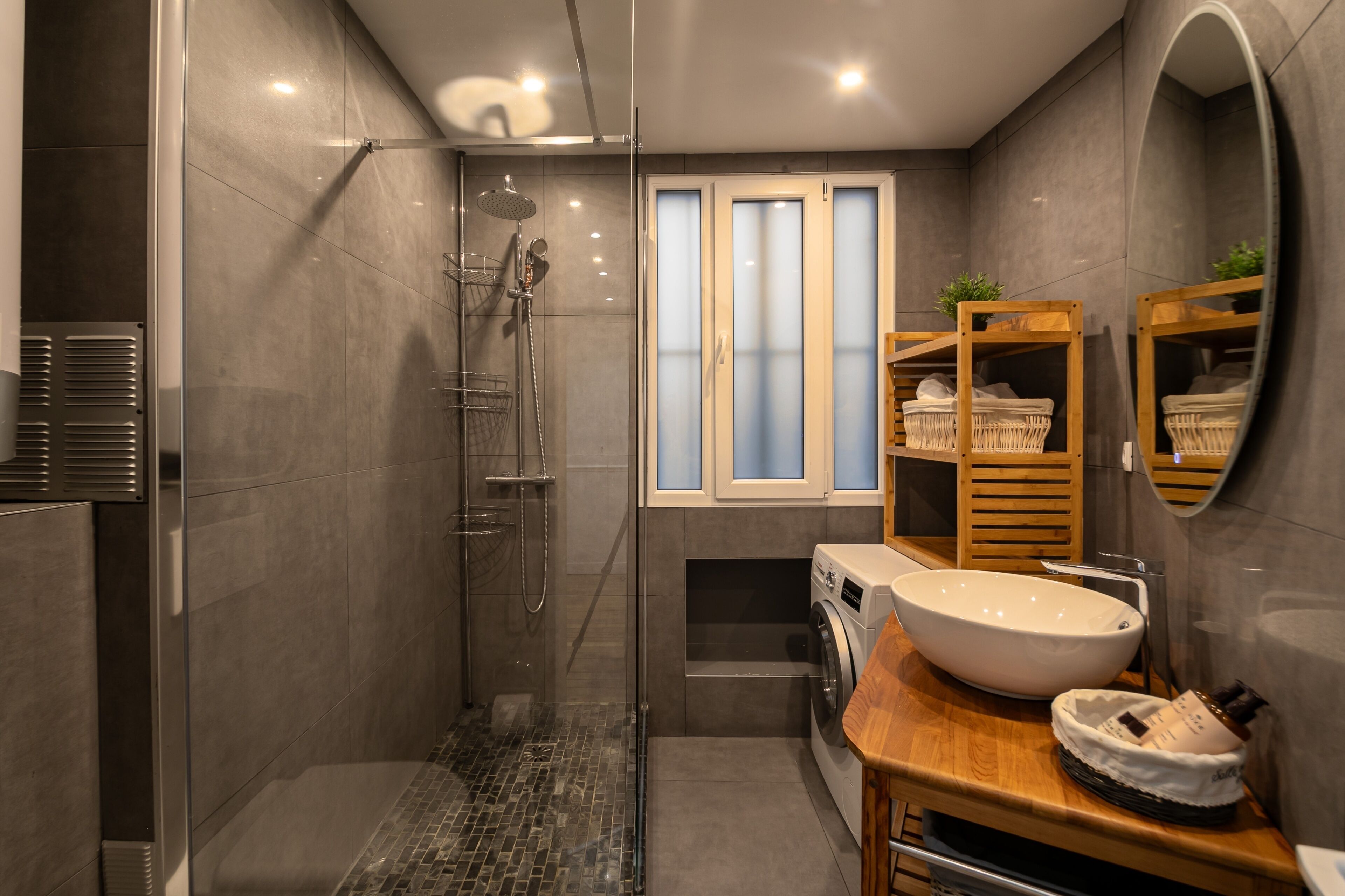 A modern bathroom with a glass-enclosed shower, a large sink, a washing machine, and a wooden vanity.