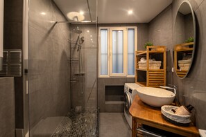 Un bany modern amb plat de dutxa envoltat de vidre, un lavabo gran, una rentadora i un tocador de fusta.