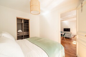 Un dormitori amb un llit, un armari, una manta verda, una làmpada penjant i una vista a una altra habitació amb un sofà i una televisió.