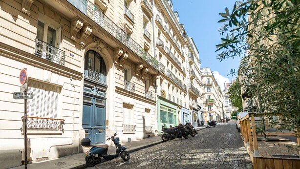 En brostensbelagt gade med historiske bygninger, en scooter parkeret på fortovet, og et skilt der angiver en parkeringsforbudszone.
