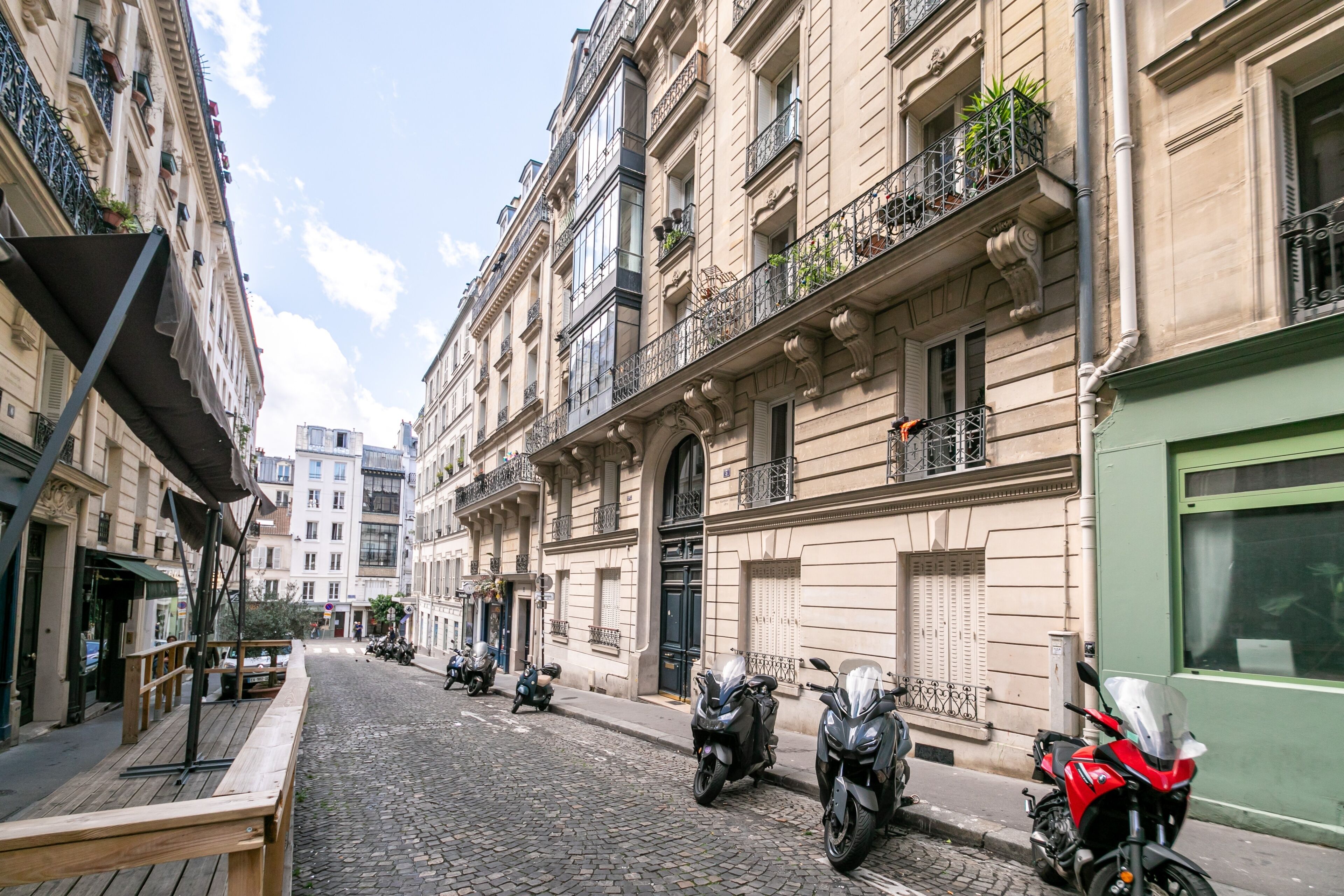 Une rue pavée bordée de bâtiments historiques, de scooters garés et d’une moto rouge.
