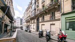 En brostensbelagt gade kantet af gamle bygninger, parkerede scootere og en rød motorcykel.