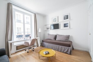 A room with a bed, a desk, a chair, a small table, and a window with curtains. - Alma Tour Eiffel - One Bedroom Apartment, Sleeps 3 (Paris)