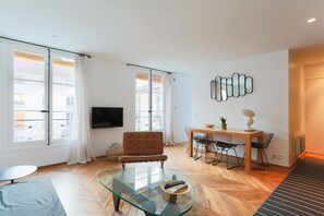 Ein modernes Wohnzimmer mit einem Glastisch, einer Couch, einem Essbereich mit Stühlen, einem an der Wand montierten Fernseher und großen Fenstern mit Vorhängen.