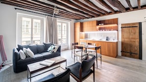 A modern living room with a sofa, coffee table, and chairs, a kitchen with wooden cabinets, and a large window with curtains. - Île Saint Louis Cosy (Paris)