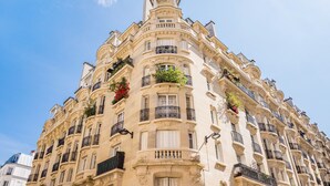 Un edifici de diversos pisos amb detalls arquitectònics ornamentals, balcons amb baranes negres i jardineres amb flors.