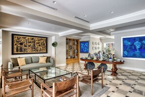 Un salon comprenant un canapé, une table basse, des fauteuils, une table avec un plateau en verre, une bibliothèque et des œuvres d’art accrochées aux murs.