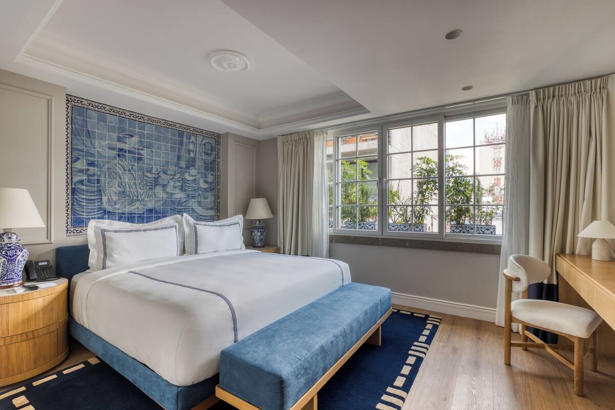 Une chambre d’hôtel avec un grand lit, un bureau, une chaise, une fenêtre avec des rideaux et un mur à motifs bleus.