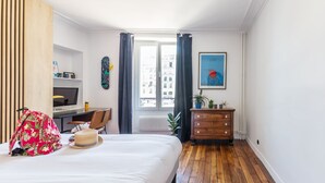 A hotel room with a bed, wooden floor, a desk with a chair, a dresser, a window with curtains, and a city view. - Canal St Martin Valmy (Paris)