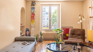 A cozy living room with a sofa, a coffee table, a bookshelf, a chair, and a window with a view of buildings. - Canal St Martin Valmy (Paris)