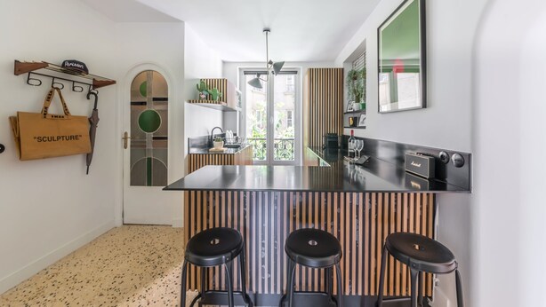 A modern kitchen with a central island, bar stools, a hanging bag labeled 'Sculpture,' and a wooden slatted wall. - Canal St Martin Valmy (Paris)