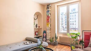 A cozy living room with a sofa, a bookshelf, a window with a view of buildings, and a radiator. - Canal St Martin Valmy (Paris)