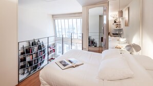 Une chambre moderne avec un grand lit, une table de chevet avec une lampe, une bibliothèque et une vue sur la ville grâce à de grandes fenêtres.