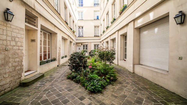 Un carrer estret amb un jardí central, paviment de pedra i edificis amb balcons i finestres.