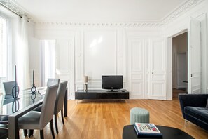 A modern living room with a glass dining table, chairs, a flat-screen TV, a sofa, and a small ottoman. - Grands Boulevards Family (Paris)