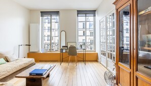A room with a wooden floor, a sofa, a table with a book, a chair, a desk with a computer, and large windows with a view of buildings. - Marais Enfants Rouges (Paris)